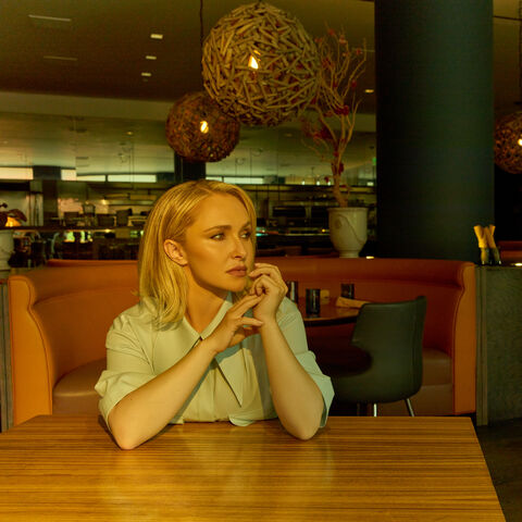 Hayden Panettiere seated alone at a restaurant table in a pale mint shirt, her chin resting on her folded hands as she gazes thoughtfully into the distance beneath warm woven pendant lights.