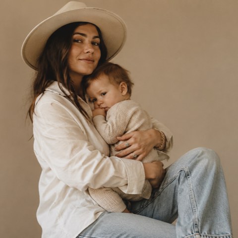 A mother in a wide-brimmed hat and white shirt holding her baby close against a warm neutral backdrop.