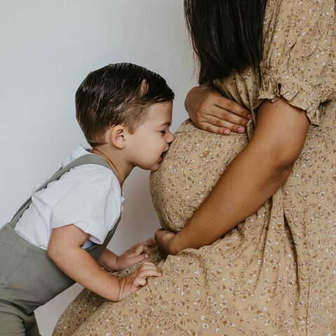 A smiling toddler in a white shirt and suspenders kisses his pregnant mother's belly, her hand resting gently on the bump.
