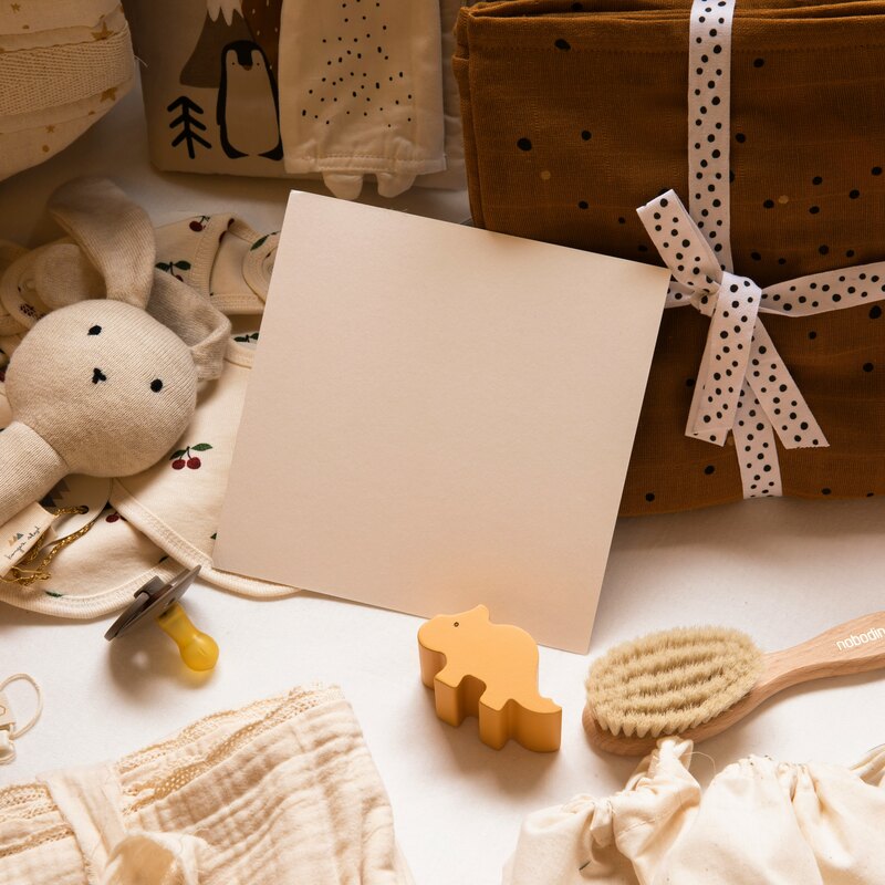 A neutral-toned flat-lay of baby items — a cream knit bunny rattle, a cherry-print onesie, a wooden brush, muslin wraps tied with polka-dot ribbon, and a blank cream card.