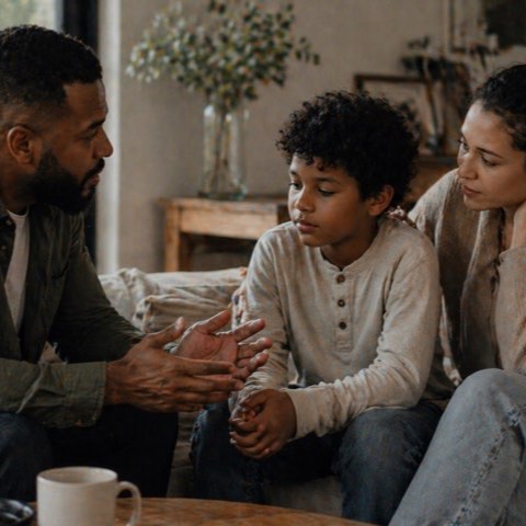 A father, mother, and young son sitting close together on a sofa, mid-conversation.